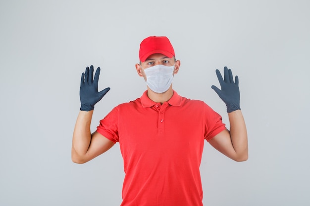 Uomo di consegna alzando le mani in uniforme rossa, mascherina medica, vista frontale guanti.