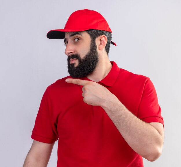 Uomo di consegna caucasico bello giovane dispiaciuto che indossa l'uniforme rossa e cappuccio che indica al lato isolato su fondo bianco