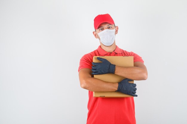 Uomo di consegna che abbraccia la scatola di cartone in uniforme rossa, mascherina medica, guanti e che sembra fiducioso