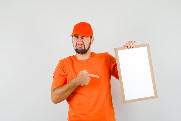 Uomo di consegna che indica al telaio vuoto in maglietta arancione, berretto e sembra infastidito. vista frontale.