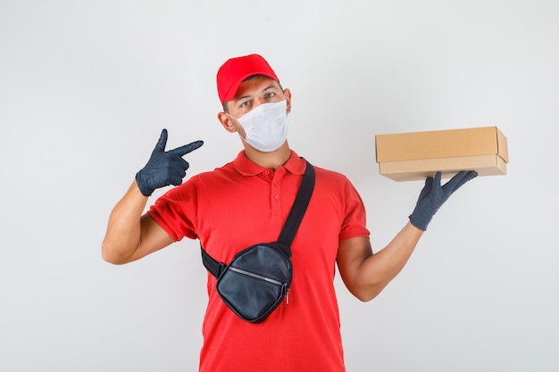 Uomo di consegna che mostra la scatola di cartone in mano in uniforme rossa, mascherina medica