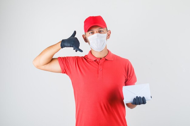 Uomo di consegna che tiene buste e che fa gesto di chiamata in uniforme rossa, mascherina medica, guanti, vista frontale.