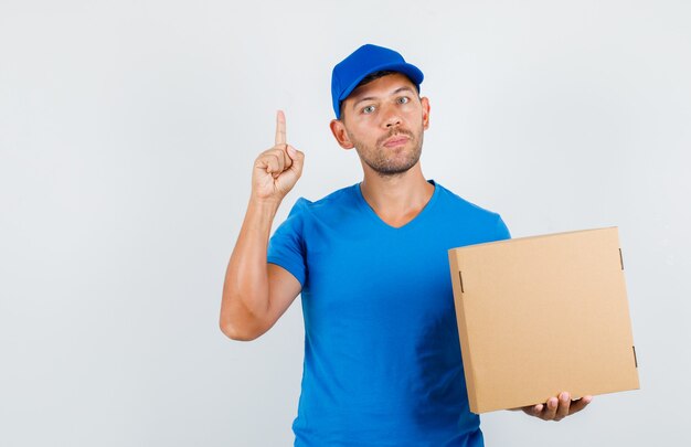 Uomo di consegna che tiene la scatola di cartone con il dito in alto in maglietta blu
