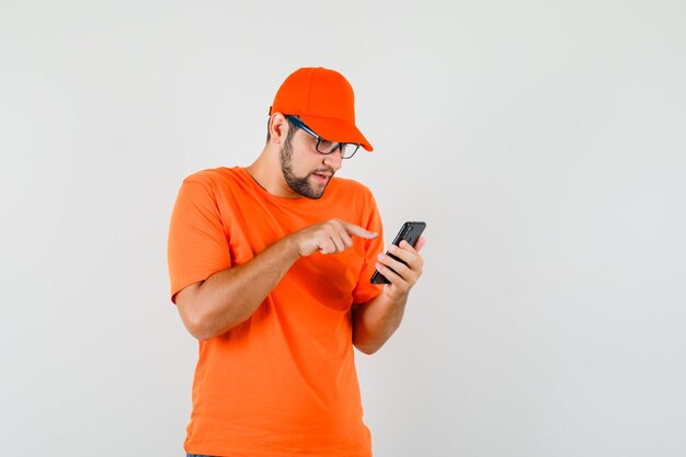 Uomo di consegna che utilizza il telefono cellulare in maglietta arancione, berretto e sembra occupato. vista frontale.