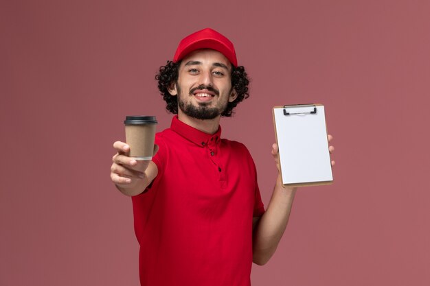 Uomo di consegna corriere maschio vista frontale in camicia rossa e mantello che tiene tazza di caffè marrone e blocco note sul lavoro maschio dipendente consegna uniforme servizio parete rosa chiaro