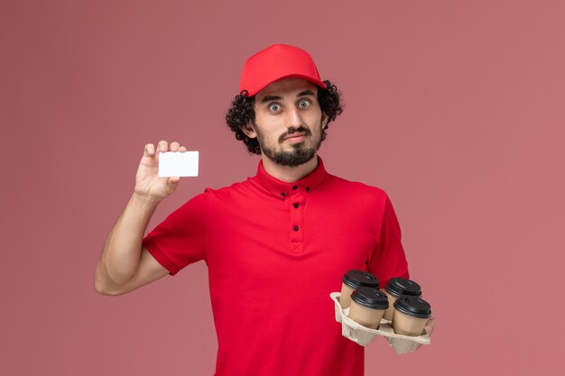 Uomo di consegna corriere maschio vista frontale in camicia rossa e mantello che tiene tazze di caffè consegna marrone con carta di plastica sul lavoro dipendente consegna servizio parete rosa