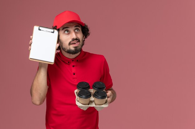 Uomo di consegna corriere maschio vista frontale in camicia rossa e mantello che tiene tazze di caffè consegna marrone con piccolo blocco note pensando sul dipendente di consegna servizio parete rosa