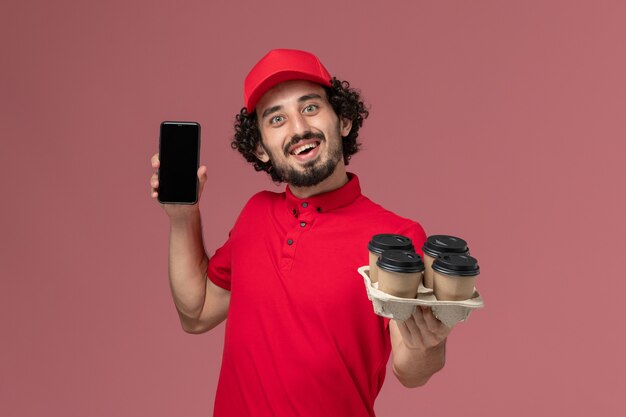 Uomo di consegna corriere maschio vista frontale in camicia rossa e mantello che tiene tazze di caffè consegna marrone con telefono sul lavoro di lavoro dipendente consegna servizio parete rosa