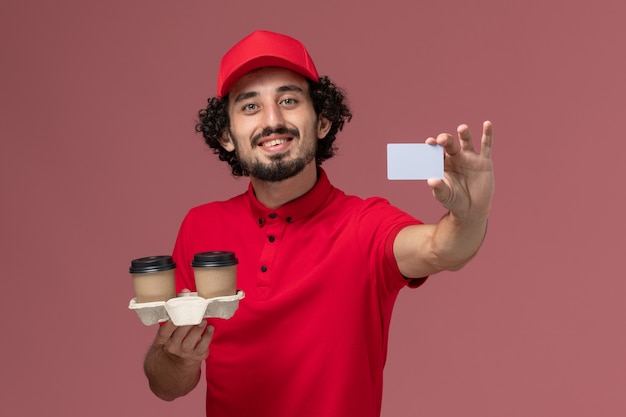 Uomo di consegna corriere maschio vista frontale in camicia rossa e mantello che tiene tazze di caffè di consegna marrone e carta sorridente sul dipendente di consegna servizio parete rosa chiaro