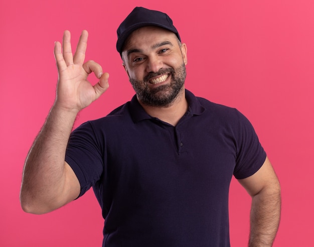 Uomo di consegna di mezza età sorridente in uniforme e cappuccio che mostra gesto giusto isolato sulla parete rosa