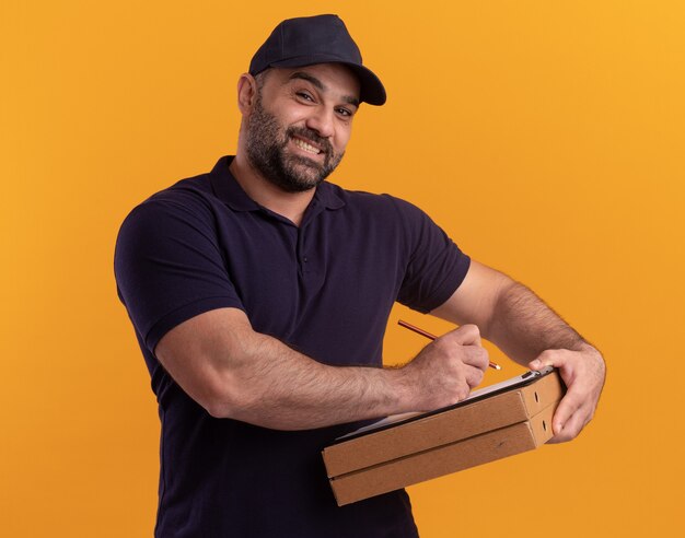 Uomo di consegna di mezza età sorridente in uniforme e cappuccio che scrive qualcosa sugli appunti sulle scatole della pizza isolate sulla parete gialla