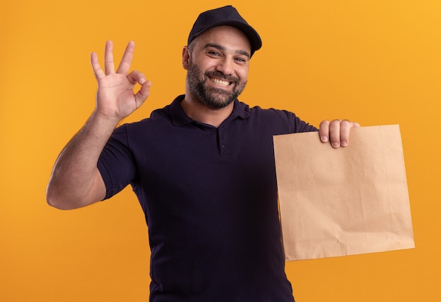 Uomo di consegna di mezza età sorridente in uniforme e cappuccio che tiene il pacchetto di cibo di carta che mostra gesto giusto isolato sulla parete gialla