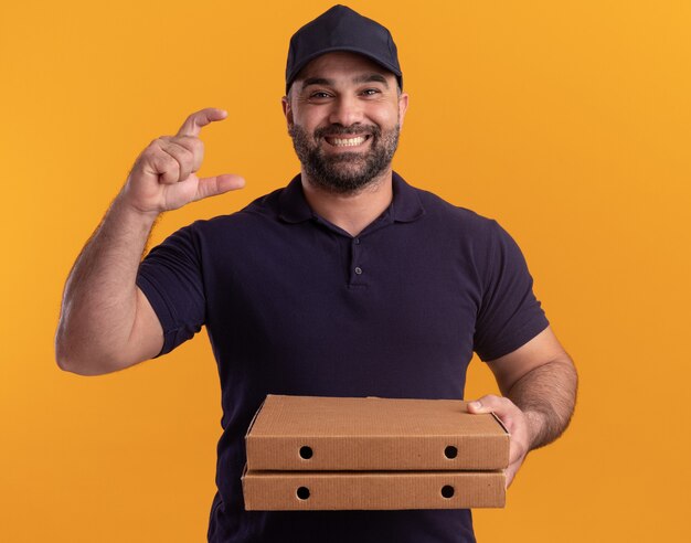 Uomo di consegna di mezza età sorridente in uniforme e cappuccio che tiene le scatole per pizza che mostrano le dimensioni isolate sulla parete gialla