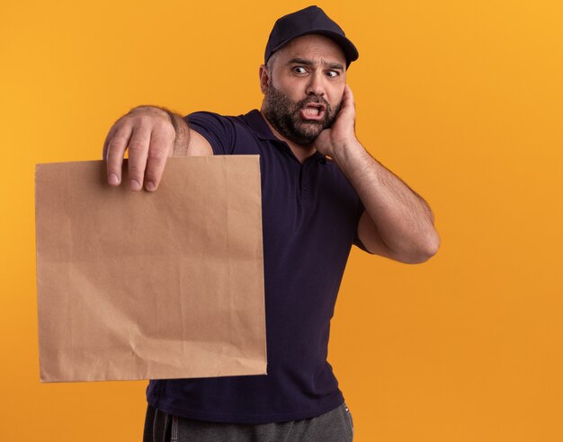 Uomo di consegna di mezza età spaventato in uniforme e cappuccio che tiene fuori il pacchetto di cibo di carta isolato sulla parete gialla