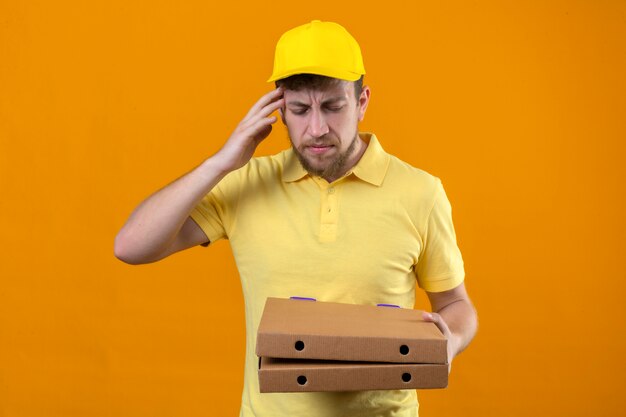 Uomo di consegna in maglietta polo gialla e cappuccio tenendo scatole per pizza in piedi con con la mano sulla testa per errore ricordare l'errore dimenticato il concetto di cattiva memoria su arancione isolato