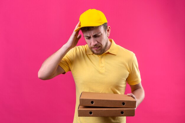 Uomo di consegna in maglietta polo gialla e cappuccio tenendo scatole per pizza in piedi con con la mano sulla testa per errore ricordare l'errore dimenticato il concetto di cattiva memoria sul rosa isolato
