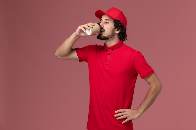 Uomo di consegna maschio corriere vista frontale in camicia rossa e mantello che beve caffè sulla parete rosa servizio uniforme consegna dipendente lavoro lavoratore lavoro maschio