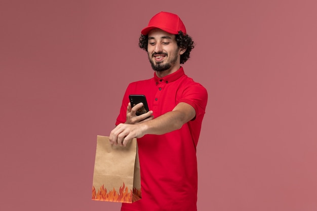 Uomo di consegna maschio del corriere di vista frontale in camicia rossa e mantello che tiene il pacchetto di cibo e il suo telefono sul dipendente di consegna del servizio di parete rosa