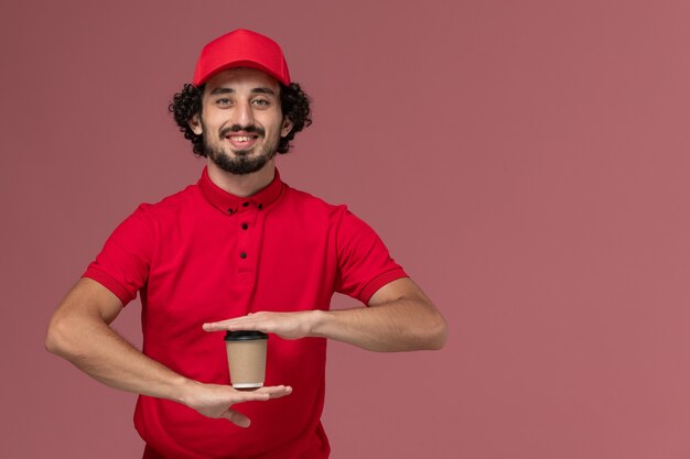 Uomo di consegna maschio del corriere di vista frontale in camicia rossa e mantello che tiene la tazza di caffè marrone di consegna sull'impiegato di consegna di servizio della parete rosa-chiaro