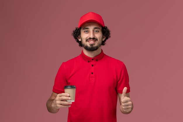Uomo di consegna maschio del corriere di vista frontale in camicia rossa e mantello che tiene la tazza di caffè marrone sul lavoro dell'operaio di lavoro dell'impiegato di consegna uniforme di servizio della parete rosa