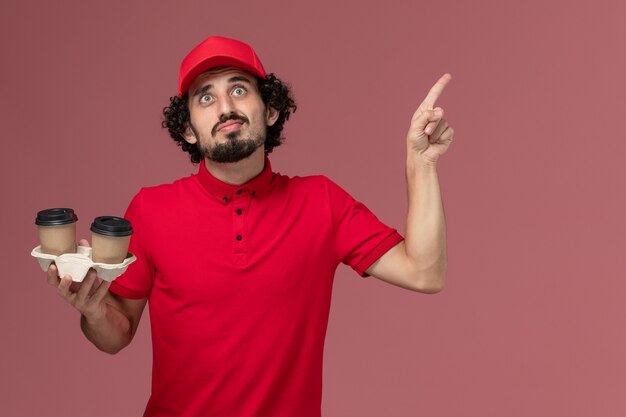 Uomo di consegna maschio del corriere di vista frontale in camicia rossa e mantello che tiene le tazze di caffè marroni di consegna che posano con il dito alzato sull'impiegato di consegna di servizio della parete rosa