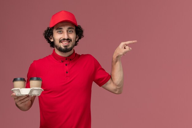 Uomo di consegna maschio del corriere di vista frontale in camicia rossa e mantello che tiene le tazze di caffè marroni di consegna sull'impiegato dell'operaio di consegna di servizio della parete rosa