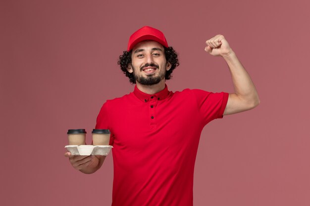 Uomo di consegna maschio del corriere di vista frontale in camicia rossa e mantello che tiene le tazze di caffè marroni di consegna sull'impiegato di consegna di servizio del lavoratore della parete rosa