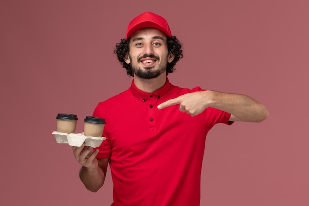 Uomo di consegna maschio del corriere di vista frontale in camicia rossa e mantello che tiene le tazze di caffè marroni di consegna sull'impiegato di consegna di servizio della parete rosa