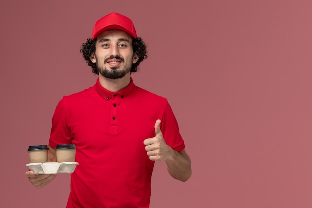 Uomo di consegna maschio del corriere di vista frontale in camicia rossa e mantello che tiene le tazze di caffè marroni di consegna sull'impiegato maschio di lavoro di consegna di servizio della parete rosa