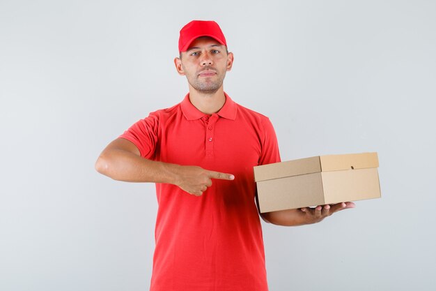 Uomo di consegna puntare il dito contro la scatola di cartone in berretto rosso e t-shirt e guardando fiducioso
