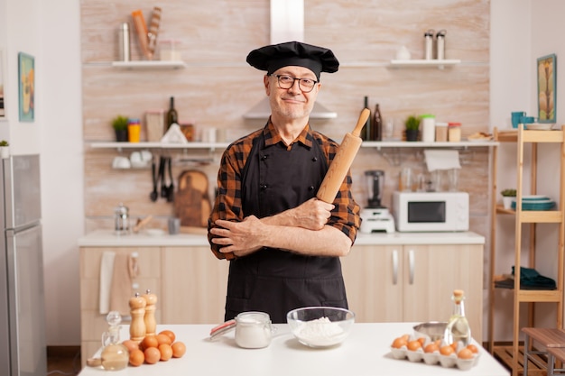 Uomo di età anziana che indossa lo chef bonete sorridente nella cucina di casa. Fornaio in pensione in uniforme da cucina che prepara gli ingredienti della pasticceria su un tavolo di legno pronto per cucinare pane, torte e pasta gustosi fatti in casa