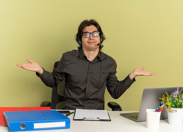 Uomo di giovane impiegato confuso sulle cuffie in vetri ottici si siede alla scrivania con strumenti di ufficio utilizzando e guardando il computer portatile