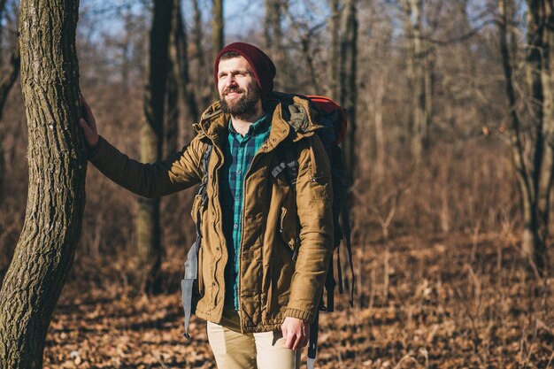 Uomo di hipster che viaggia con lo zaino nella foresta di autunno che indossa giacca calda, cappello, turista attivo, esplorando la natura nella stagione fredda