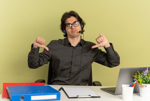 Uomo di lavoratore di ufficio infastidito giovane sulle cuffie in vetri ottici si siede alla scrivania con strumenti di ufficio utilizzando il pollice verso il basso del computer portatile isolato su priorità bassa verde con lo spazio della copia