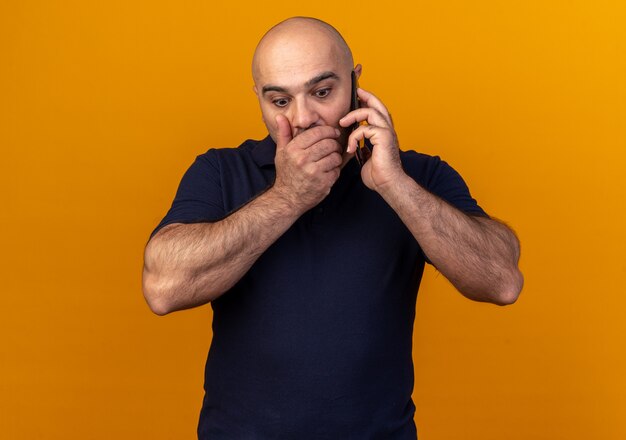 Uomo di mezza età casual interessato che parla al telefono tenendo la mano sulla bocca guardando in basso isolato sul muro arancione orange