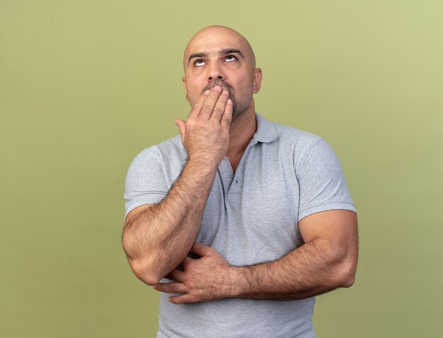 Uomo di mezza età casuale confuso che tiene la mano sulla bocca che guarda in su isolato sul muro verde oliva