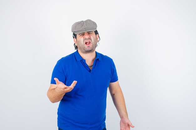 Uomo di mezza età che allunga la mano in modo interrogativo in t-shirt polo, cappello edera e sembra arrabbiato