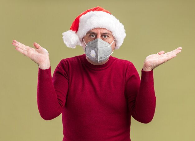 Uomo di mezza età che indossa il cappello di Babbo Natale che indossa la maschera protettiva per il viso che guarda l'obbiettivo confuso con le braccia alzate in piedi su sfondo verde