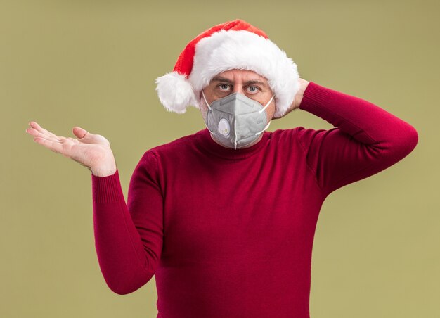 uomo di mezza età che indossa il cappello di Babbo Natale che indossa la maschera protettiva per il viso guardando la fotocamera confuso con il braccio alzato in piedi su sfondo verde