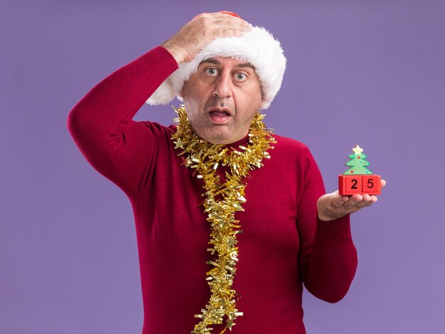 Uomo di mezza età che indossa il cappello di Babbo Natale con orpelli intorno al collo tenendo i cubi giocattolo con data venticinque guardando la telecamera preoccupato e confuso con la mano sulla sua testa in piedi su sfondo viola
