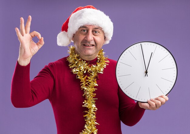 uomo di mezza età che indossa il cappello di Babbo Natale con orpelli intorno al collo tenendo l'orologio da parete guardando la fotocamera sorridente mostrando segno ok in piedi su sfondo viola