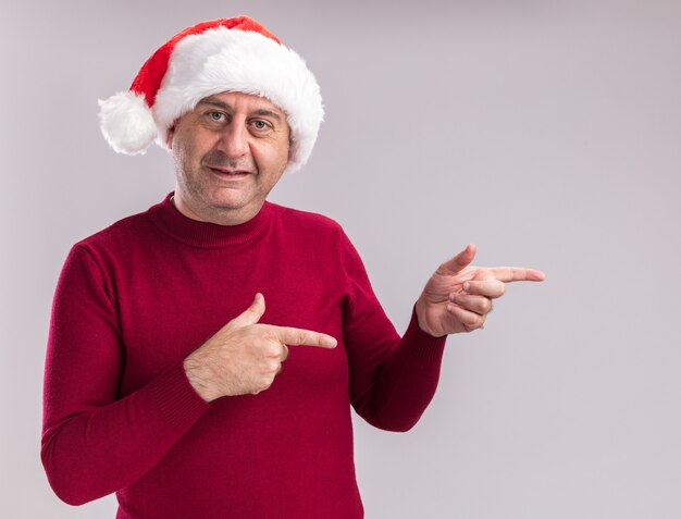 uomo di mezza età che indossa il cappello di Babbo Natale guardando la fotocamera con il sorriso sul viso che punta con figners indice al lato in piedi su sfondo bianco