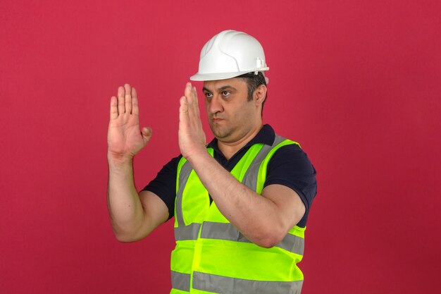 Uomo di mezza età che indossa la maglia gialla da costruzione e la direzione del casco di sicurezza che gesturing con le mani che mostrano le dimensioni sopra la parete rosa isolata