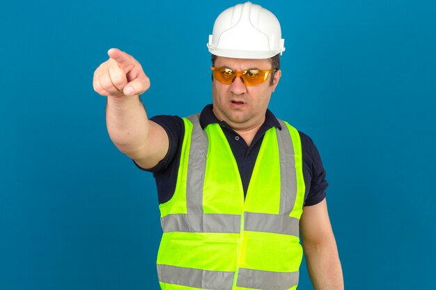 Uomo di mezza età che indossa maglia gialla costruzione e casco di sicurezza che punta a qualcosa di avanti con la faccia seria sopra isolato muro blu