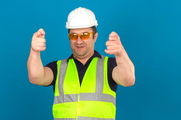 Uomo di mezza età che indossa maglia gialla di costruzione e casco di sicurezza e che puntava il dito indice sorridendo isolato muro blu