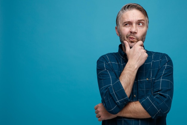 Uomo di mezza età con capelli grigi in camicia di colore scuro che guarda da parte perplesso tenendo la mano sul mento pensando in piedi su sfondo blu