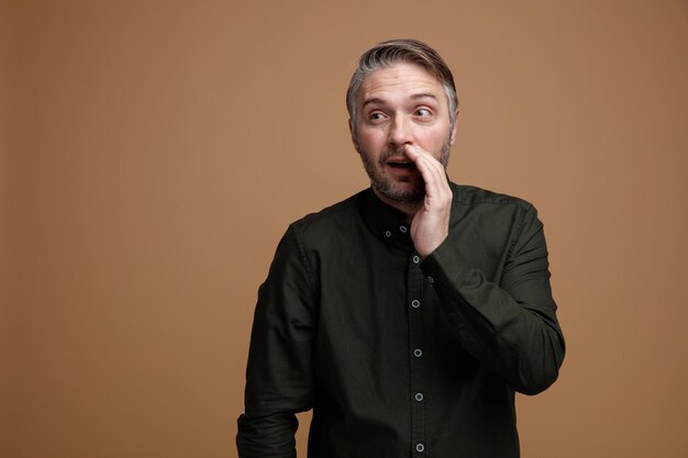 Uomo di mezza età con capelli grigi in camicia di colore scuro che guarda da parte raccontando pettegolezzi che copre la bocca con la mano in piedi su sfondo marrone