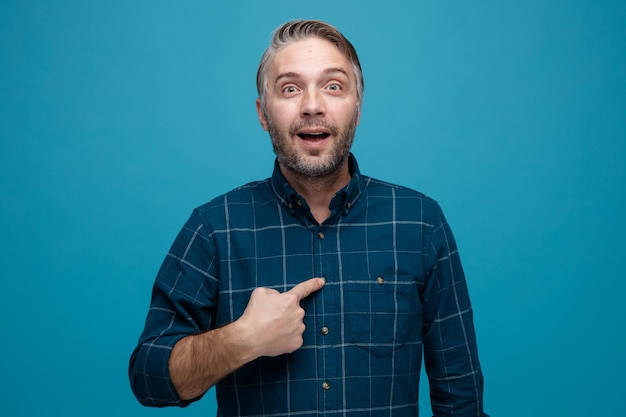 Uomo di mezza età con capelli grigi in camicia di colore scuro che guarda la fotocamera sorpreso puntando con il dito indice su se stesso in piedi su sfondo blu