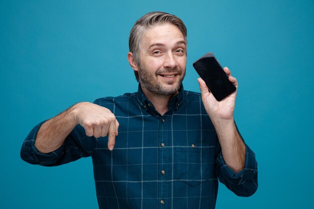 Uomo di mezza età con capelli grigi in camicia di colore scuro che mostra lo smartphone guardando la fotocamera sorridente con la faccia felice che punta con il dito indice verso il basso in piedi su sfondo blu