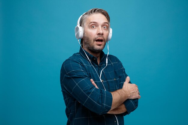Uomo di mezza età con capelli grigi in camicia di colore scuro con le cuffie che guardano la fotocamera stupito e sorpreso con le braccia incrociate sul petto in piedi su sfondo blu